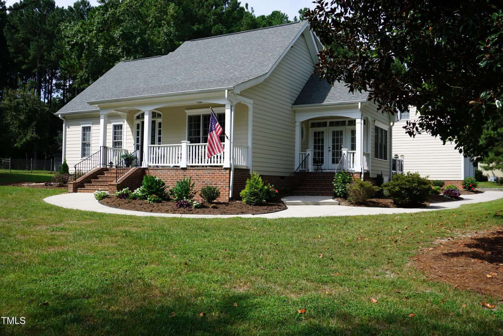 A single-story, light-colored house with a large front porch, white railings, brick steps, and an American flag; surrounded by green lawn, shrubs, and trees on a sunny day.