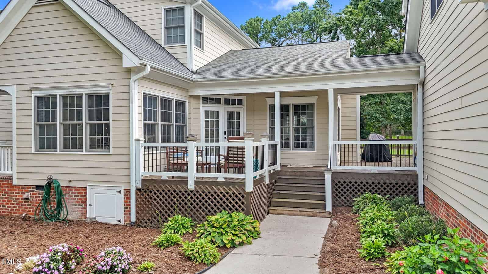 A beige house with white trim features a back patio with outdoor dining furniture, steps leading up to a deck, lattice skirting, and landscaped flower beds along a concrete walkway.
