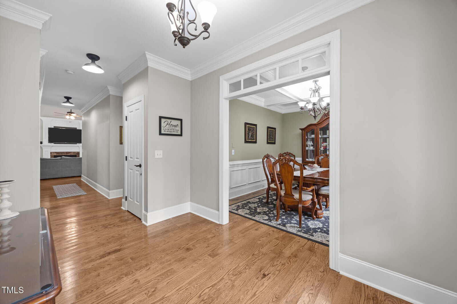 A hallway with hardwood floors and beige walls leads to a dining room with a wooden table and chairs, a china cabinet, and wall art. Chandeliers hang from the ceiling, and crown molding decorates the space.