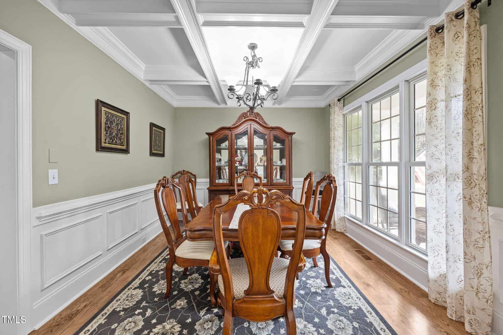Traditional dining room with a wooden table, six chairs, a china cabinet, chandelier, decorative wall panels, two framed pictures, a floral rug, and large windows with patterned curtains letting in natural light.