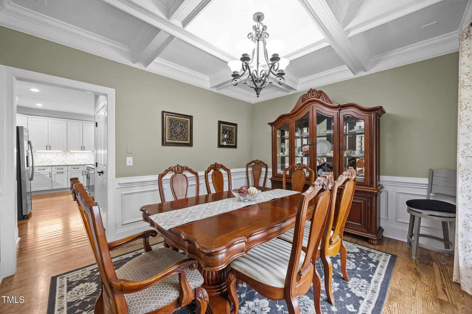 Formal dining room with coffered ceiling, chandelier, wooden table with six chairs, china cabinet, and decorative wall art. Hardwood floors, area rug, and view into a white kitchen through an open doorway.