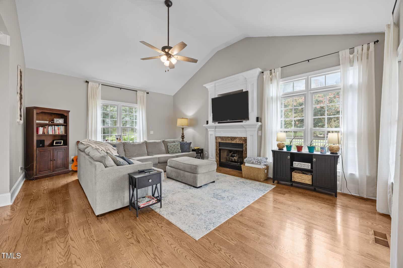A bright living room with hardwood floors, a gray sectional sofa, fireplace with mantel, TV, two windows with white curtains, a ceiling fan, a bookshelf, and a black console table with lamps and baskets.