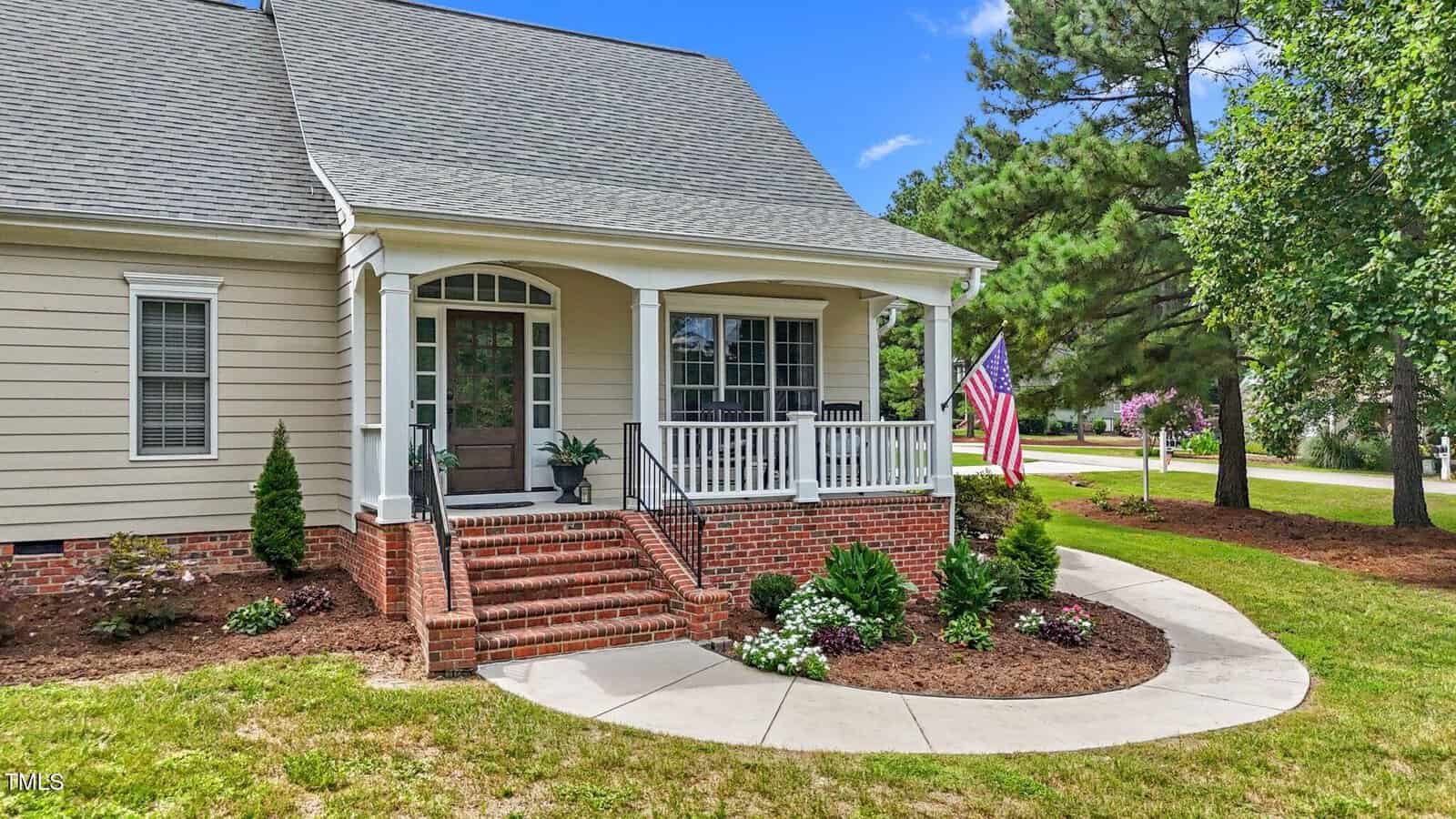 A beige house with a covered front porch, white railings, brick steps, and landscaped garden beds. An American flag hangs by the porch, and trees line the green lawn under a bright blue sky.