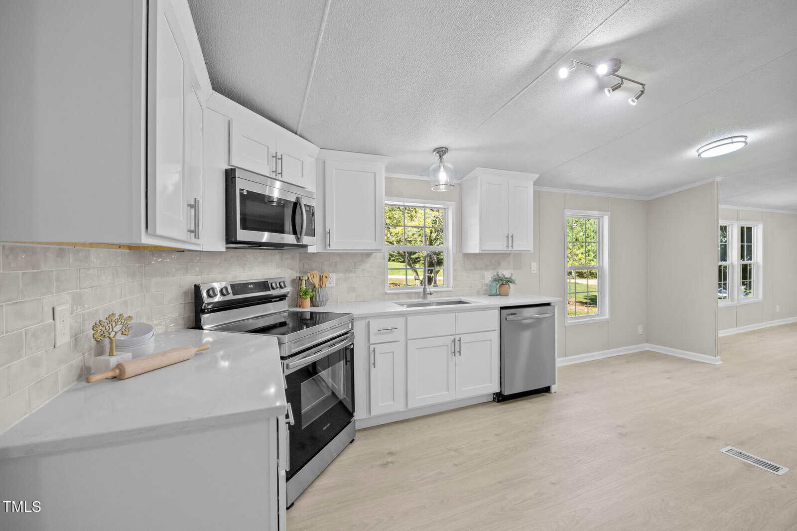Modern kitchen with white cabinets, stainless steel appliances, a large window above the sink, light wood flooring, and neutral walls. The kitchen opens into a bright living space with large windows.