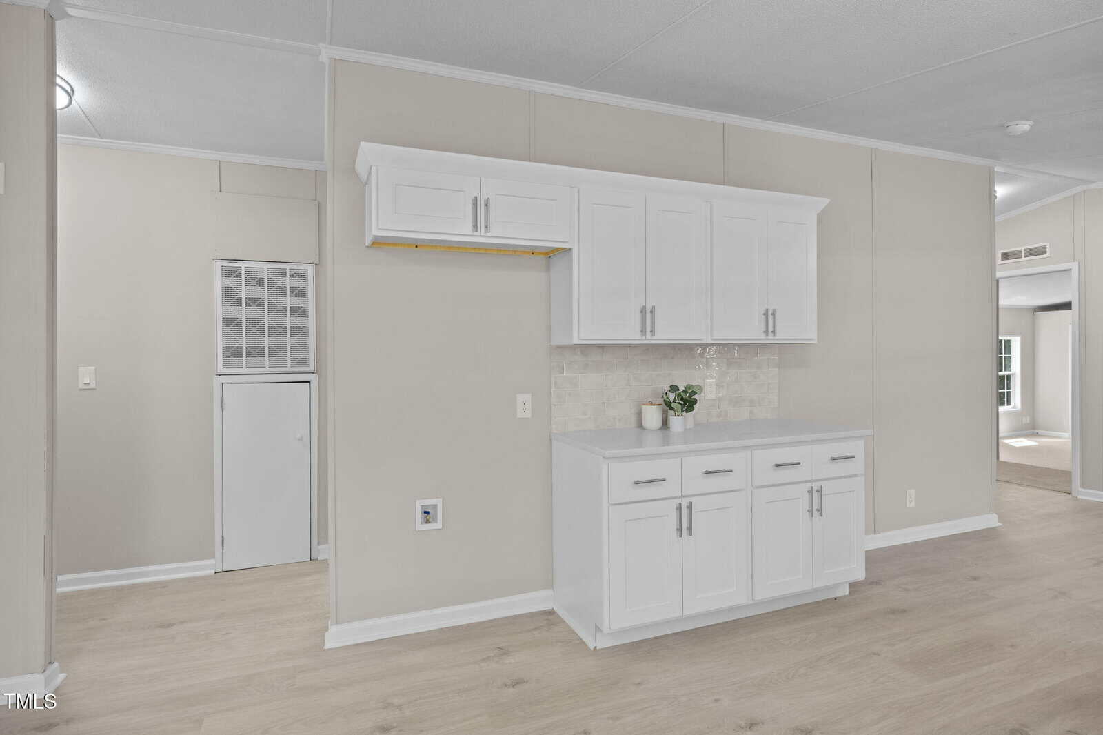 A section of a modern interior with light wood floors, beige walls, white upper and lower cabinets with silver handles, a small countertop, a potted plant, and a vented utility area on the left.