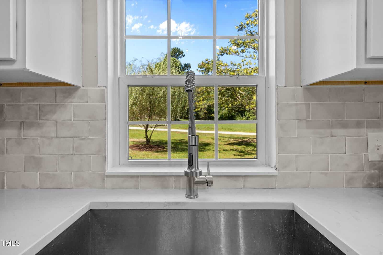 A stainless steel kitchen sink with a modern faucet is set against a white tile backsplash beneath a window, which shows a clear blue sky and green trees outside.