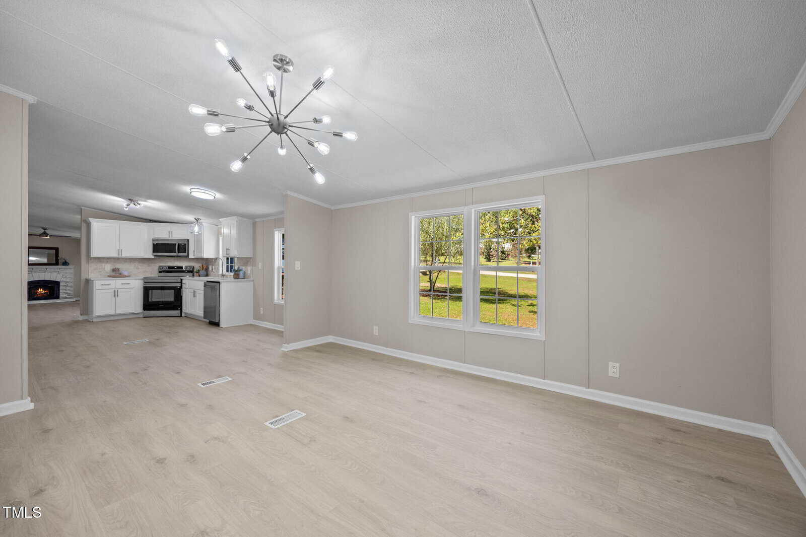 A modern, unfurnished living and kitchen area with light wood floors, beige walls, a large window overlooking a grassy yard, and a contemporary ceiling light fixture. The kitchen has white cabinets and stainless steel appliances.