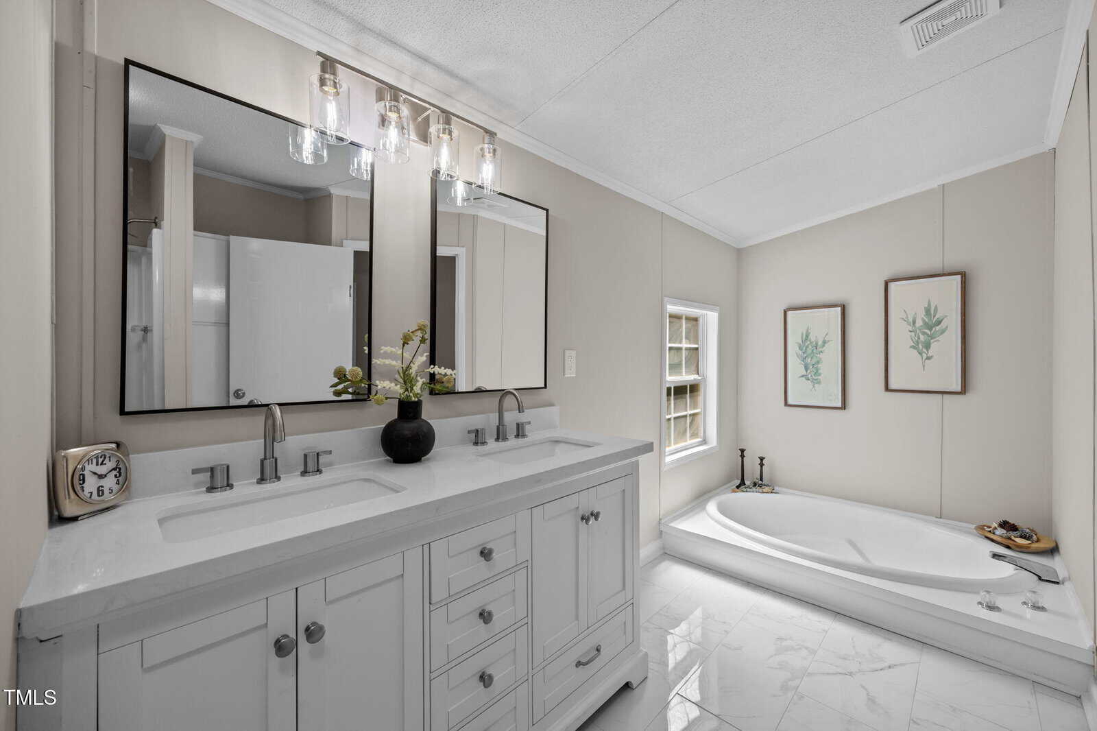 Modern bathroom with a double-sink vanity, two mirrors, and a countertop clock and vase. There is a bathtub by a window, two botanical art prints on the wall, and white tile flooring.