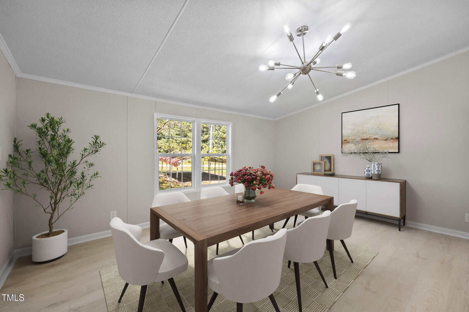 A dining room with light walls and floors, a wooden table with six white chairs, a modern chandelier, a sideboard with decor and artwork, a potted plant in the corner, and large windows letting in natural light.