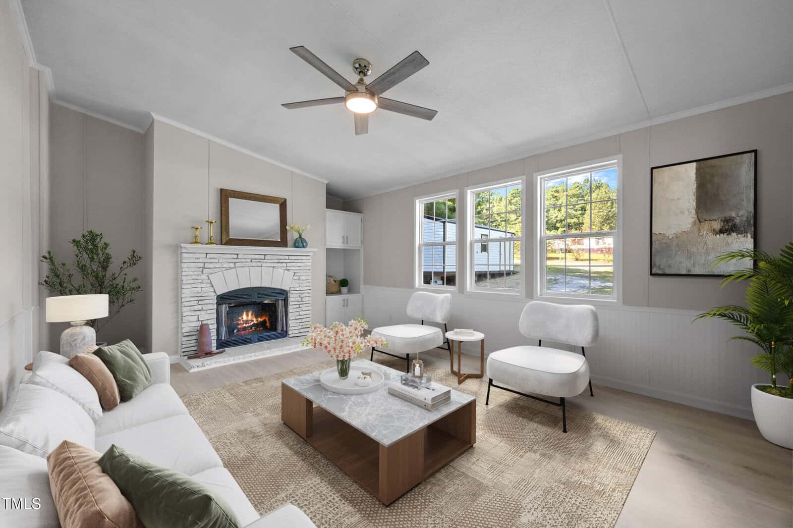 A living room with a white sofa, two white armchairs, a coffee table, a fireplace with a white brick surround, houseplants, large windows, neutral walls, and a ceiling fan with a light.
