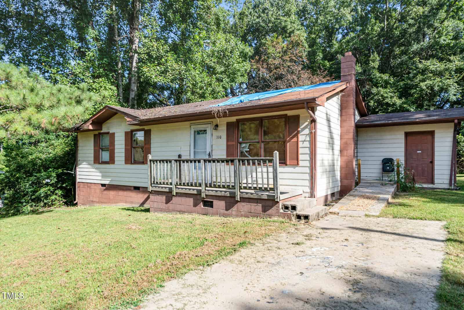 A single-story beige house with brown trim and a small front porch sits at 110 Lynnbank Estates Road, Kittrell. Part of the roof is covered with a blue tarp. The grassy yard features trees, greenery, and a concrete driveway.