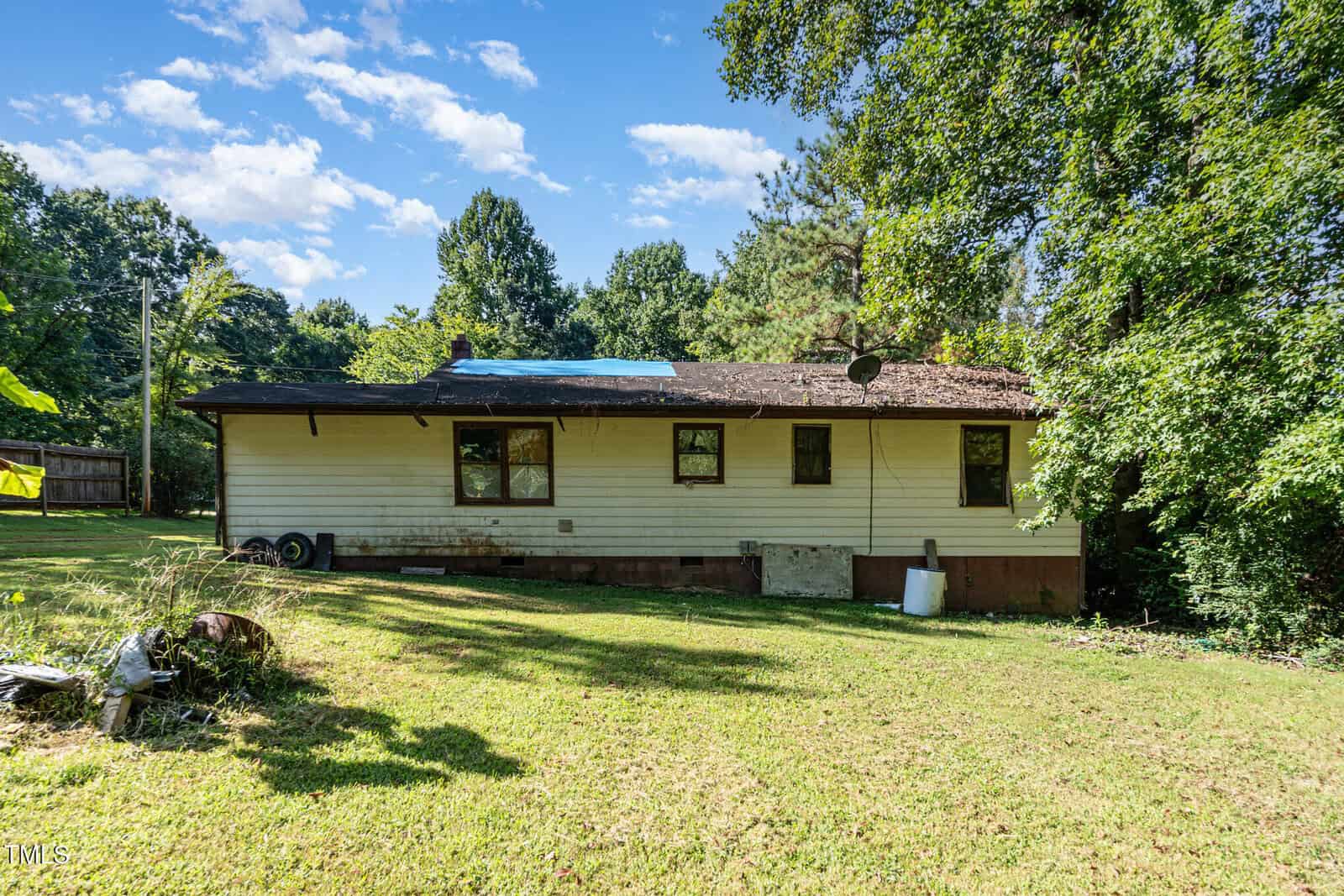 A single-story house with white siding and a brown roof is shown. There is a satellite dish on the roof and a blue tarp covering part of the shingles. The yard is grassy with trees and bushes in the background.