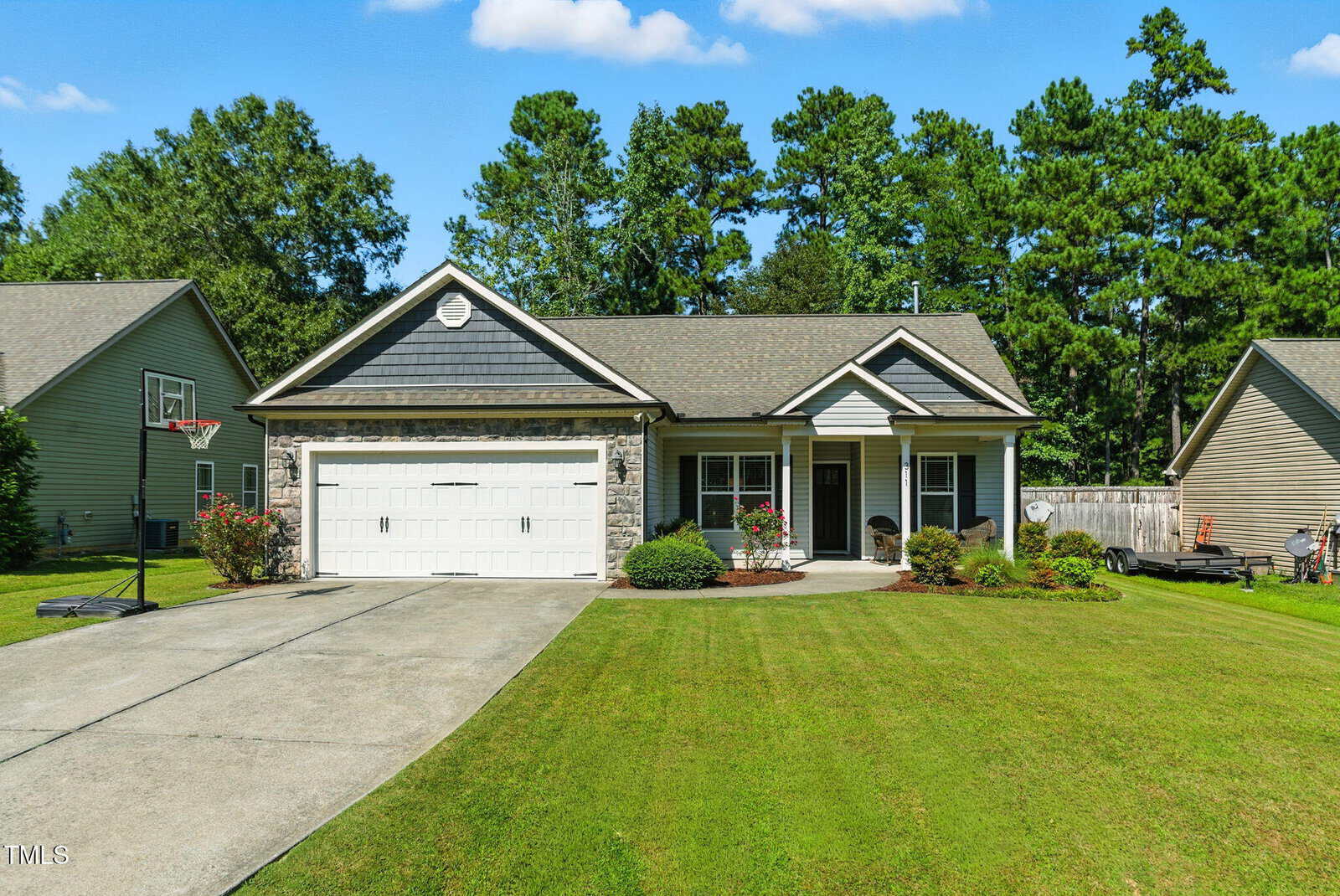 A single-story house at 311 Coachmans Trail in Stem features gray siding, a two-car garage, a covered front porch, and a well-maintained lawn with bushes, flowers by the entrance, and trees in the background under a blue sky.