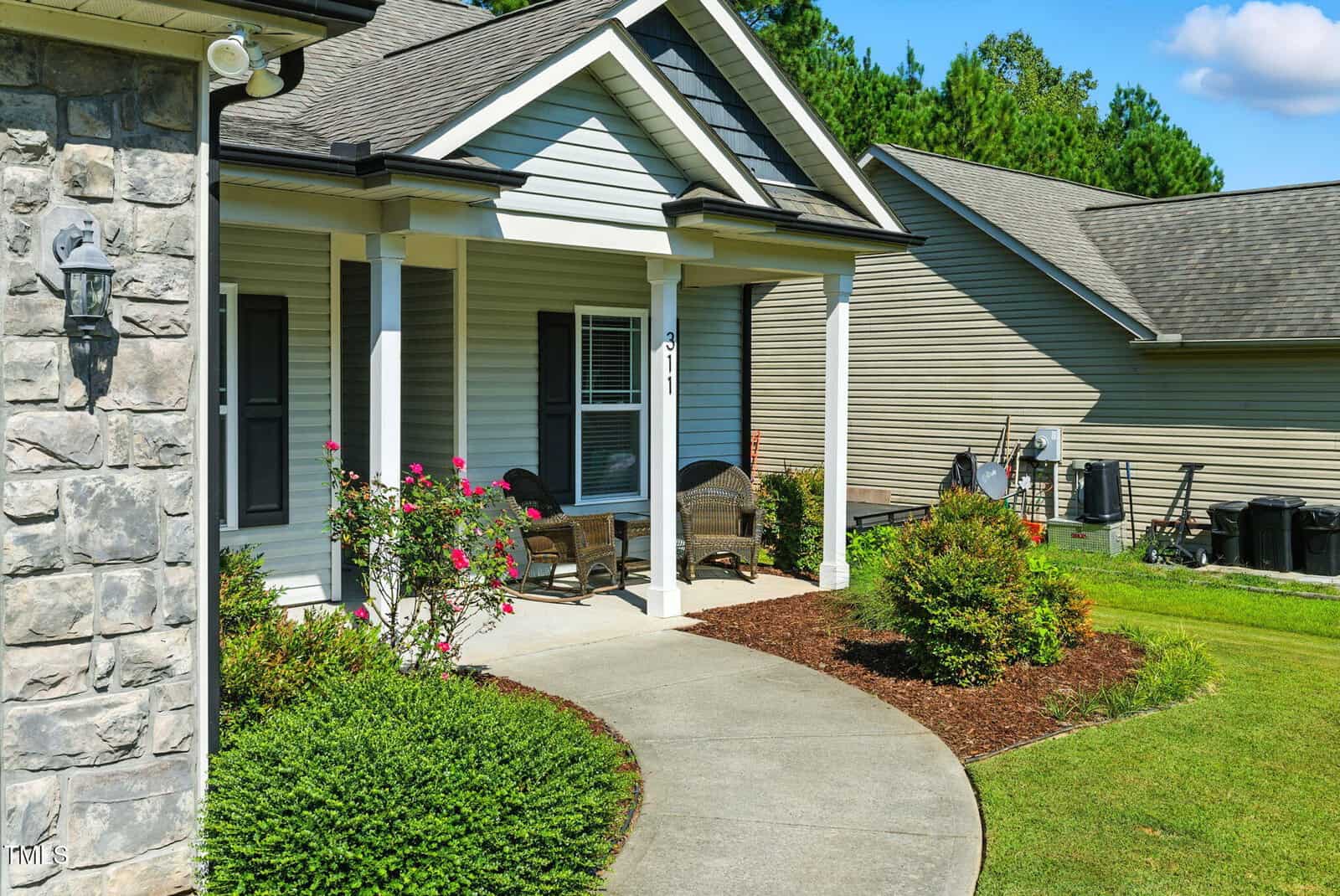A curved concrete walkway leads to a covered front porch with wicker chairs and a small table. The house has stone and siding exterior, shrubs, and a well-maintained lawn with a neighboring house visible.