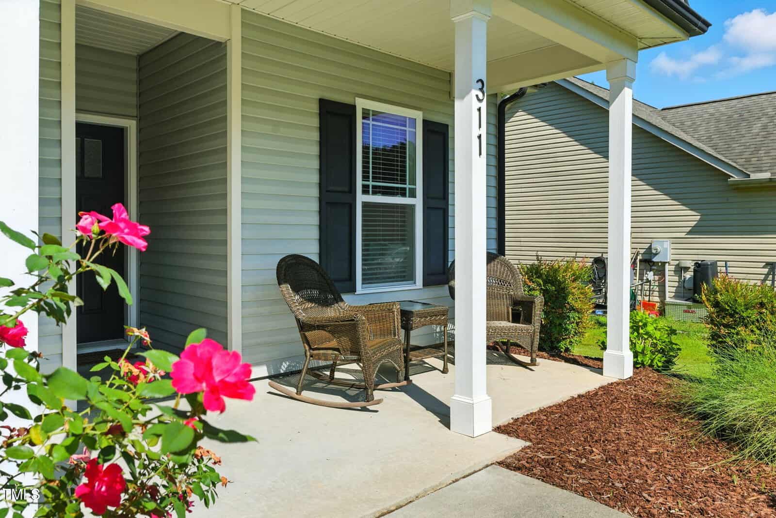 A small front porch with two wicker chairs and a matching table sits outside a gray house with white trim. Pink flowers and green shrubs border the porch. The house number 311 is visible on a porch post.