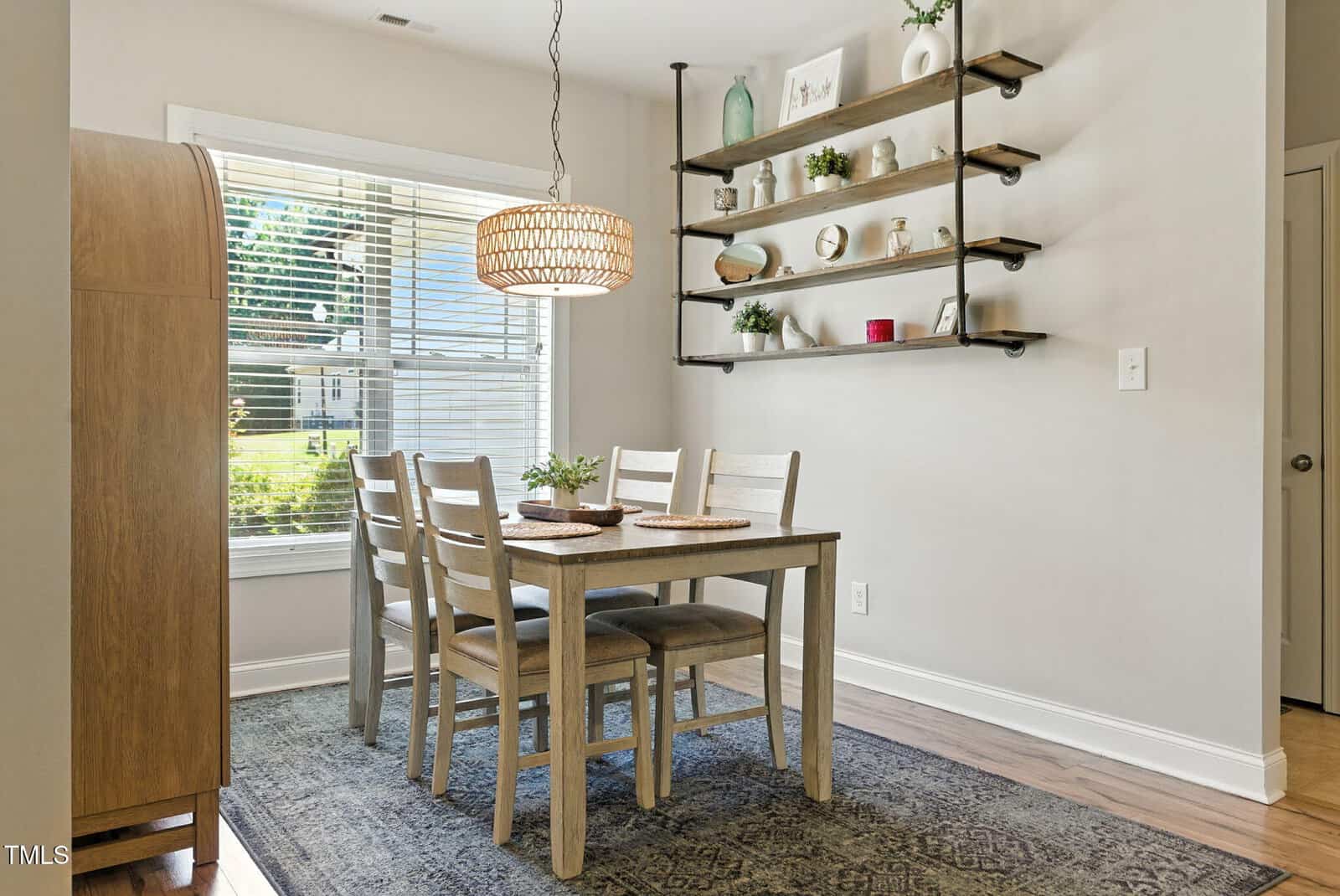A small dining area with a wooden table set for four, cushioned chairs, a patterned rug, a hanging woven light fixture, wall-mounted shelves with decor, and a large window letting in natural light.