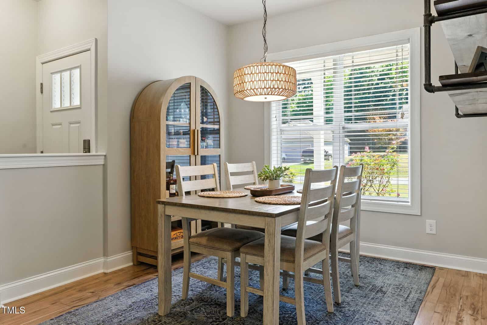A dining area with a wooden table set for four, matching chairs, a woven pendant light, a wooden cabinet, a window with blinds, and a blue rug on a hardwood floor.
