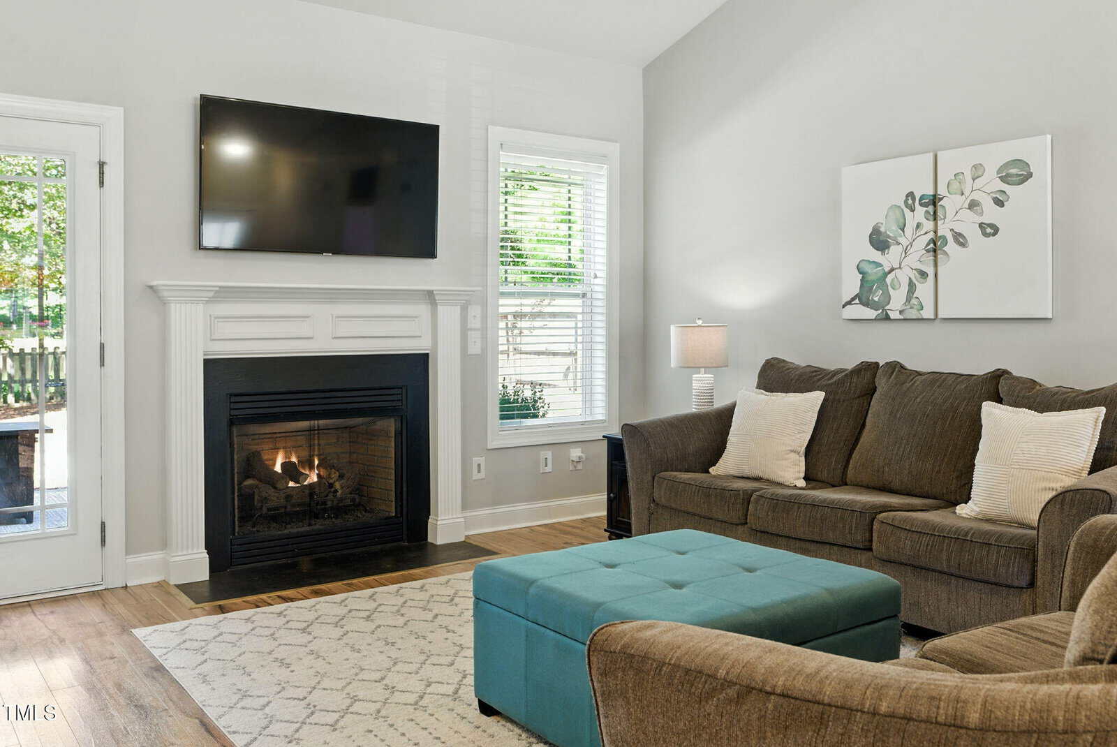 A living room with a brown sofa, a turquoise ottoman, a lit fireplace, a flat-screen TV mounted above the mantel, a patterned rug, a side table with a lamp, and two windows with white blinds.