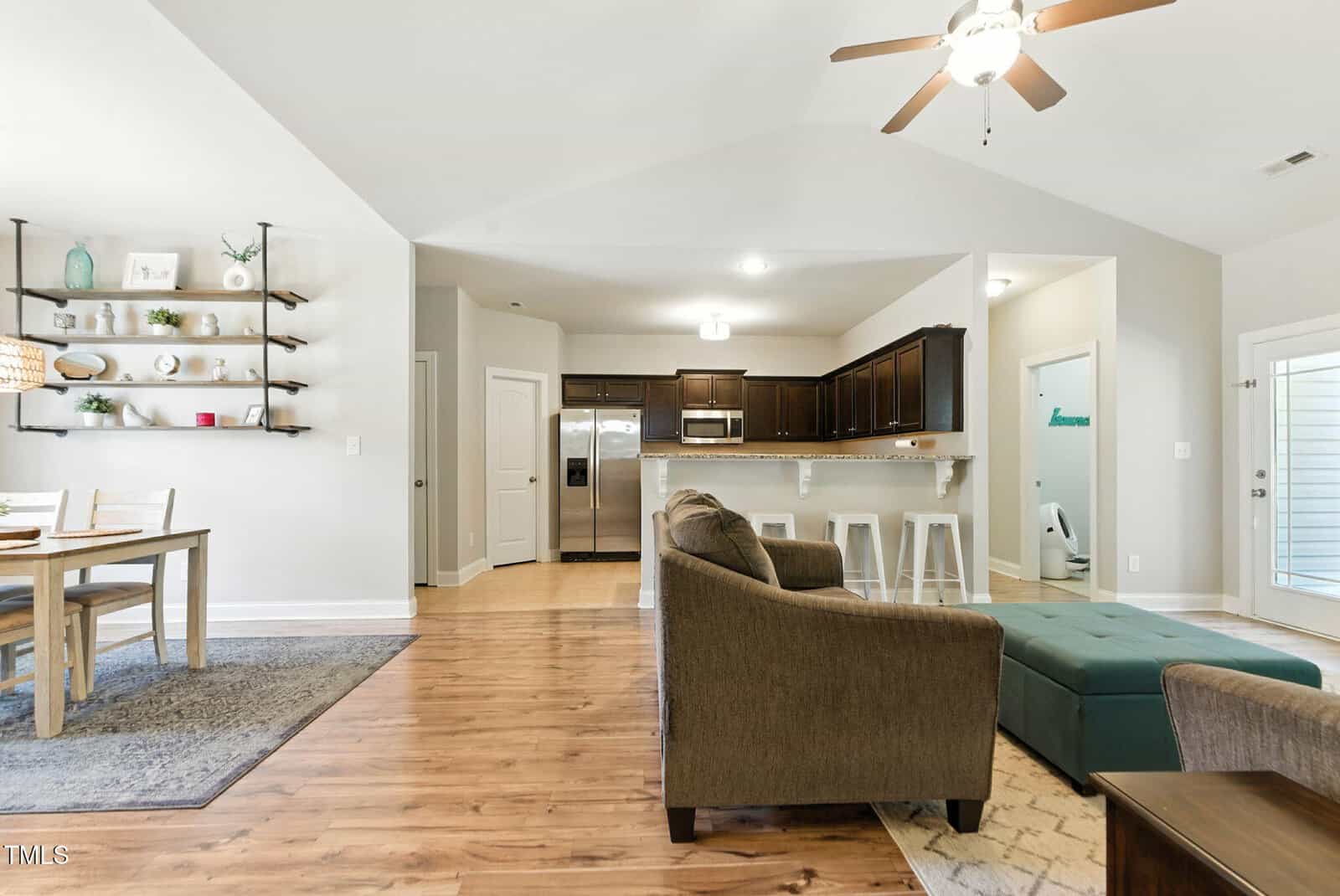 Open-concept living area with wood floors; includes a dining table, a kitchen with dark cabinets and stainless steel appliances, a breakfast bar with stools, and a living room with a sofa and ottoman. Ceiling fan above.