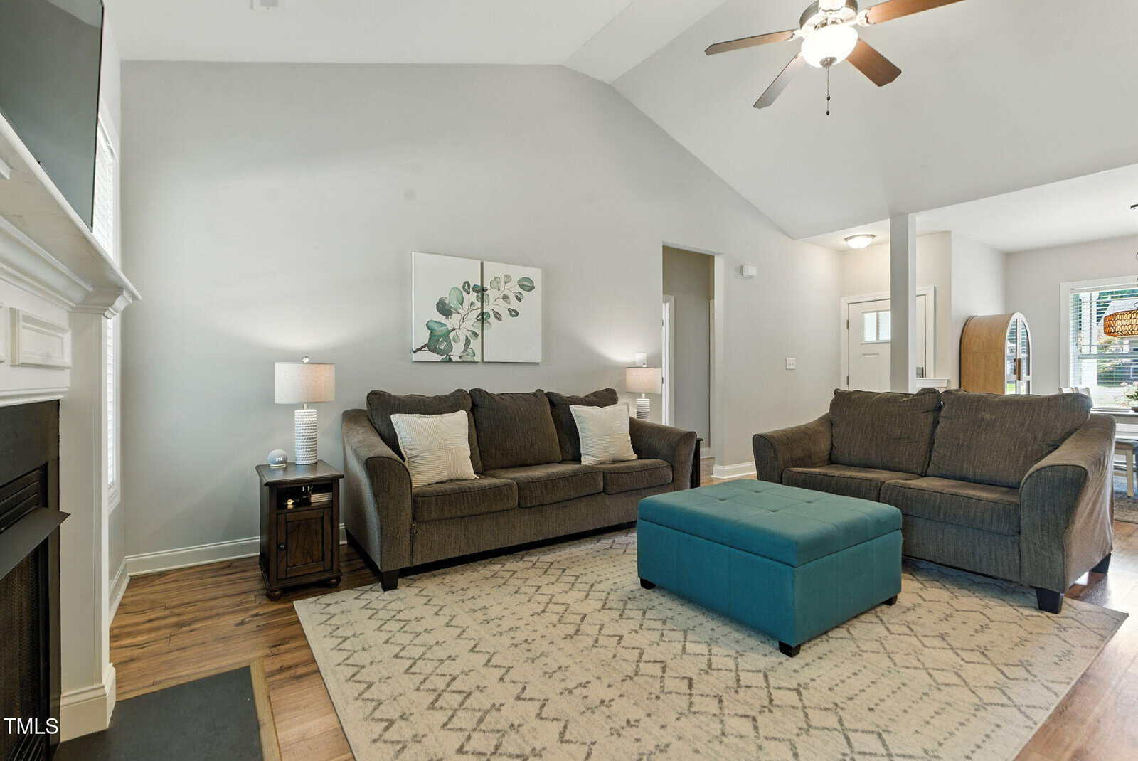 A living room with two dark sofas, a turquoise ottoman, a patterned rug, a side table with a lamp, and a ceiling fan. A piece of wall art hangs above the sofa, and the room opens to a dining area and entryway.