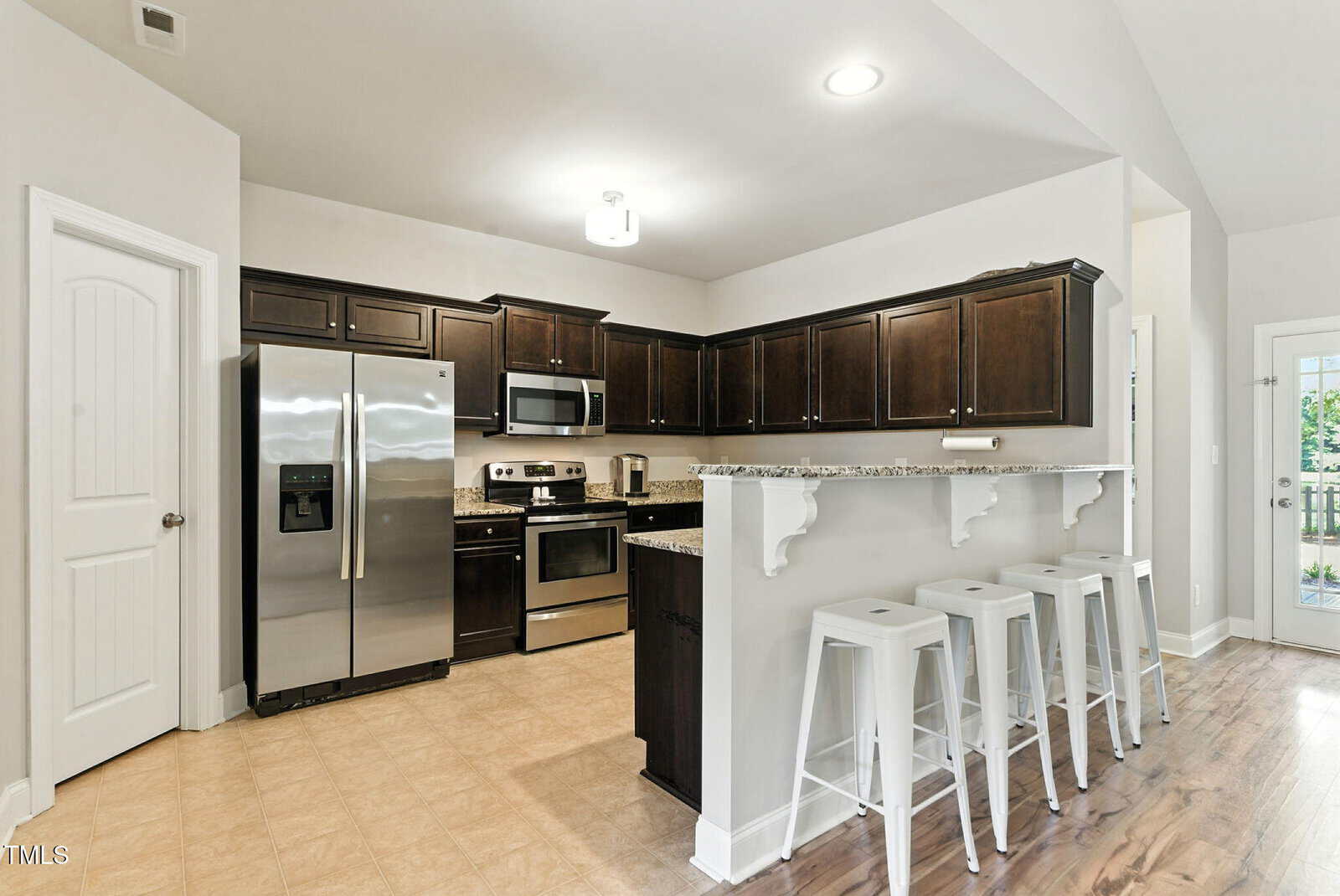 Modern kitchen with stainless steel appliances, dark wood cabinets, granite countertops, and a breakfast bar with three white bar stools. The room has light tile flooring and a white door to the left.