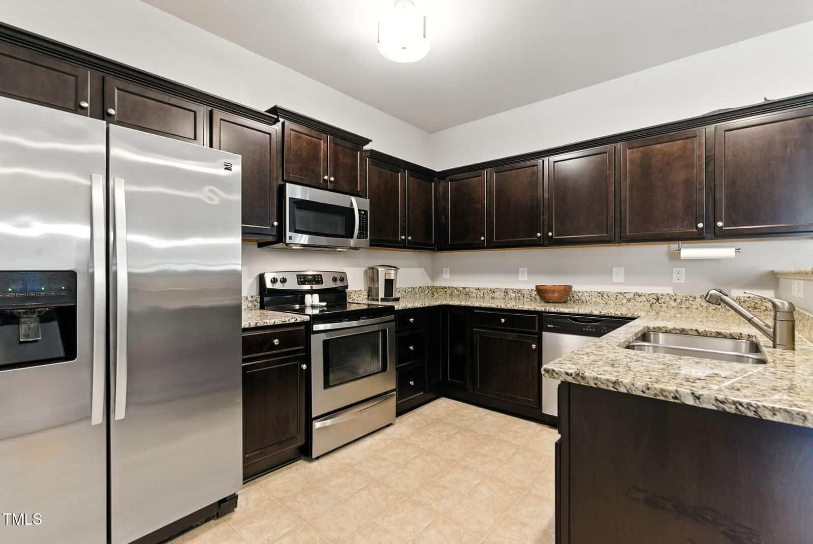 A modern kitchen featuring dark wooden cabinets, stainless steel appliances including a refrigerator, oven, and microwave, granite countertops, a double sink, and beige tile flooring.