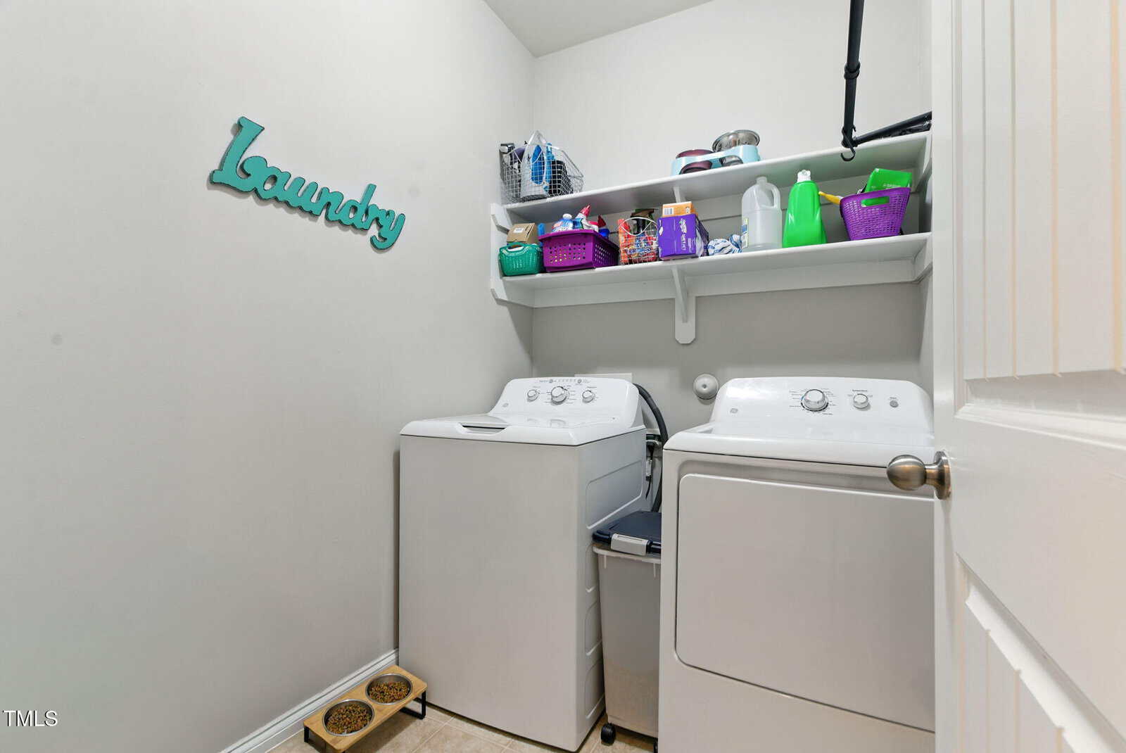 A laundry room with a washer and dryer, white shelves above holding baskets, detergent, and supplies. “Laundry” decor hangs on the wall, and there are pet food bowls on the floor.