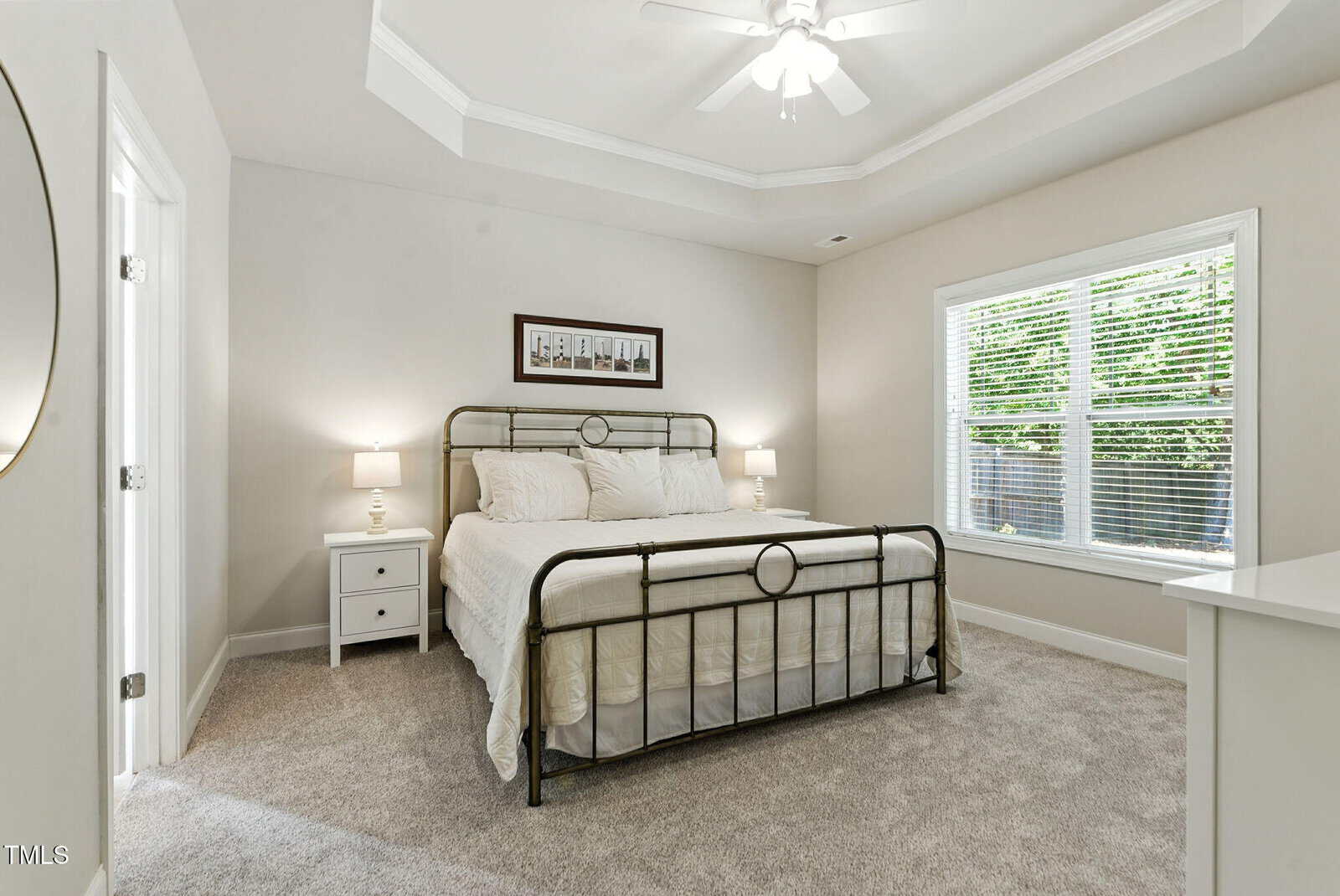 Bright bedroom with a metal bedframe, white bedding, two nightstands with lamps, carpeted floor, ceiling fan, large window with blinds, and framed photos above the bed.