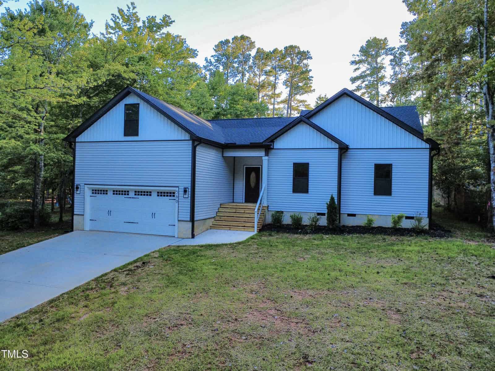 Charming single-story house at 560 Shawnee Drive, Louisburg, featuring white siding with black trim, an attached two-car garage, front steps leading to a covered entrance, and surrounded by mature trees and lush grass.
