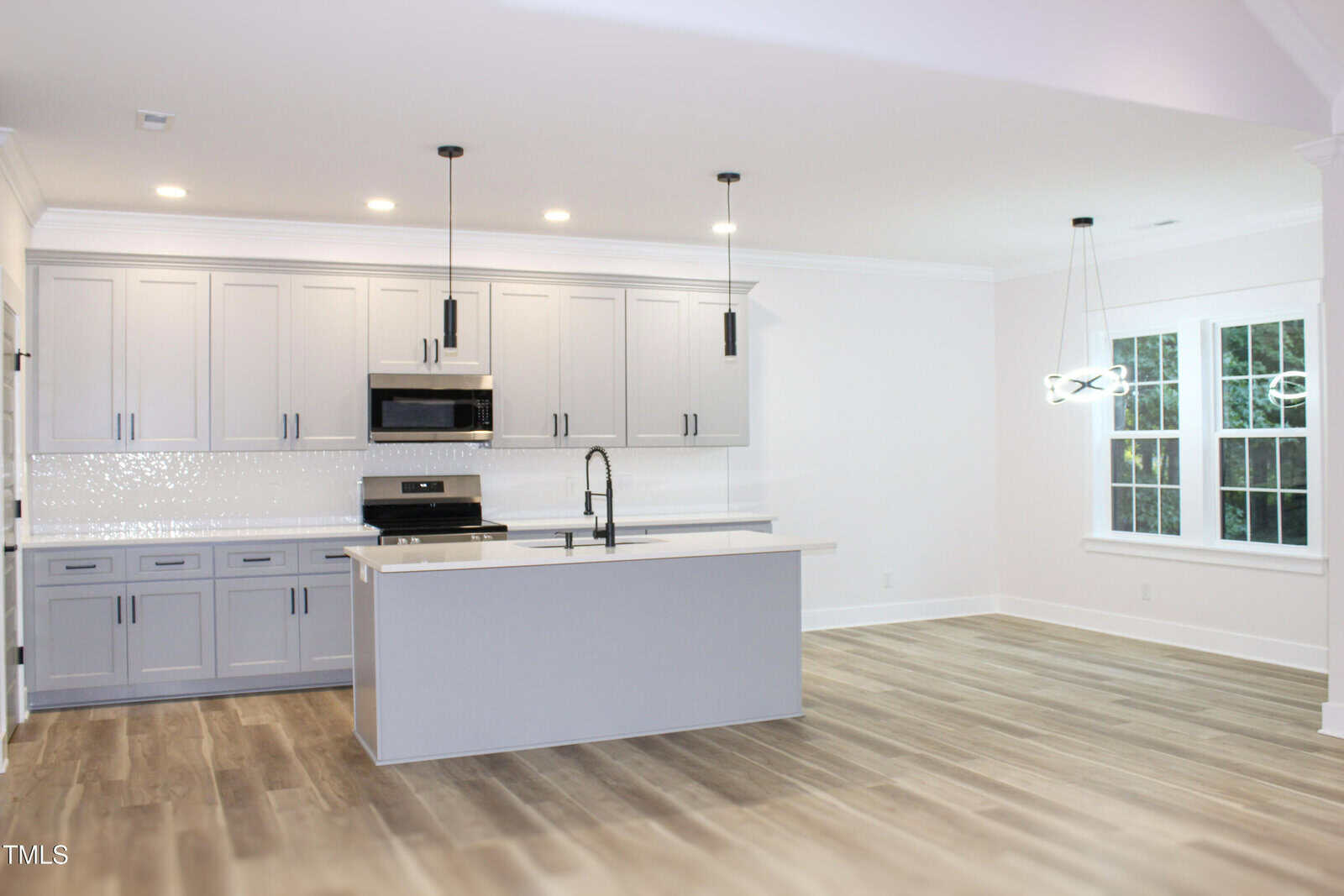Modern kitchen with light gray cabinets, island with sink, stainless steel appliances, pendant lights, white tile backsplash, and wood-look flooring; dining area with a window and modern chandelier.