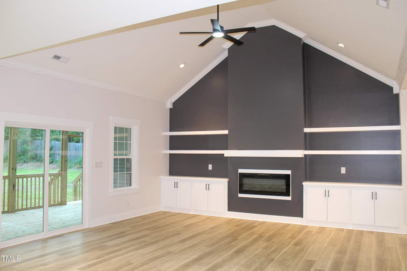 A modern living room with light wood floors, a dark accent wall featuring a built-in fireplace and cabinets, large windows, and a glass door leading to a wooden deck outside. Ceiling has a black fan and recessed lighting.