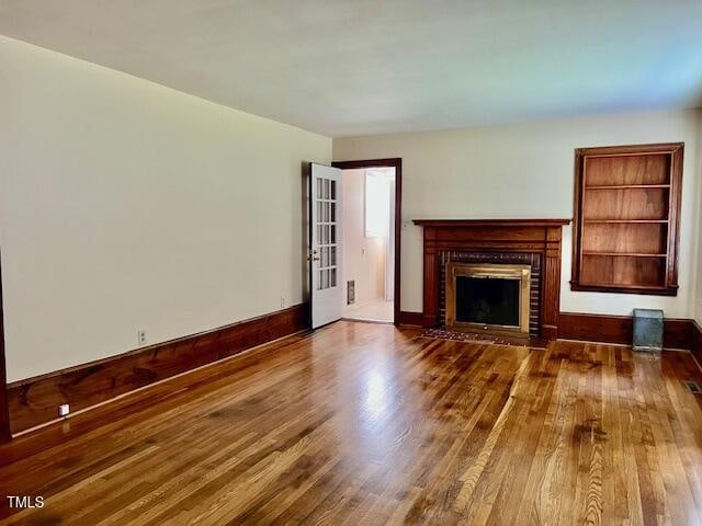 Empty living room with polished hardwood floors, a white wall, wooden trim, a fireplace with a wooden mantel, a window, and an open door leading to another room.