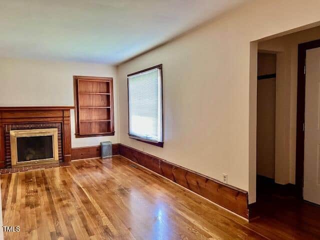 A bright, empty living room with hardwood floors, a fireplace with wood trim, built-in shelves, a large window with blinds, and cream-colored walls.