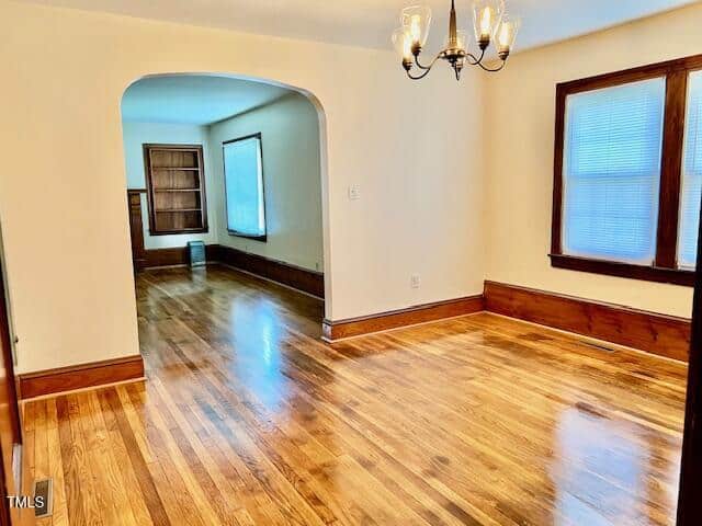 Bright dining room with hardwood floors, wood trim, chandelier, and large windows. An arched doorway leads to an adjoining room with more windows and matching hardwood floors.