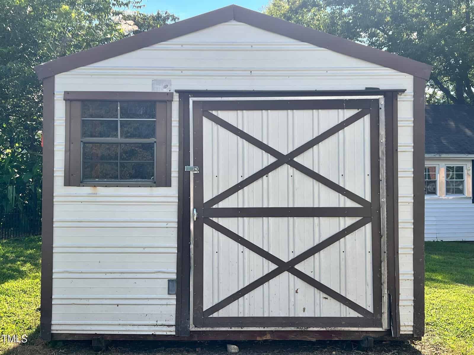 A white shed with brown trim and a large front door featuring an "X" design, a window with metal bars on the left, and grass and trees in the background.