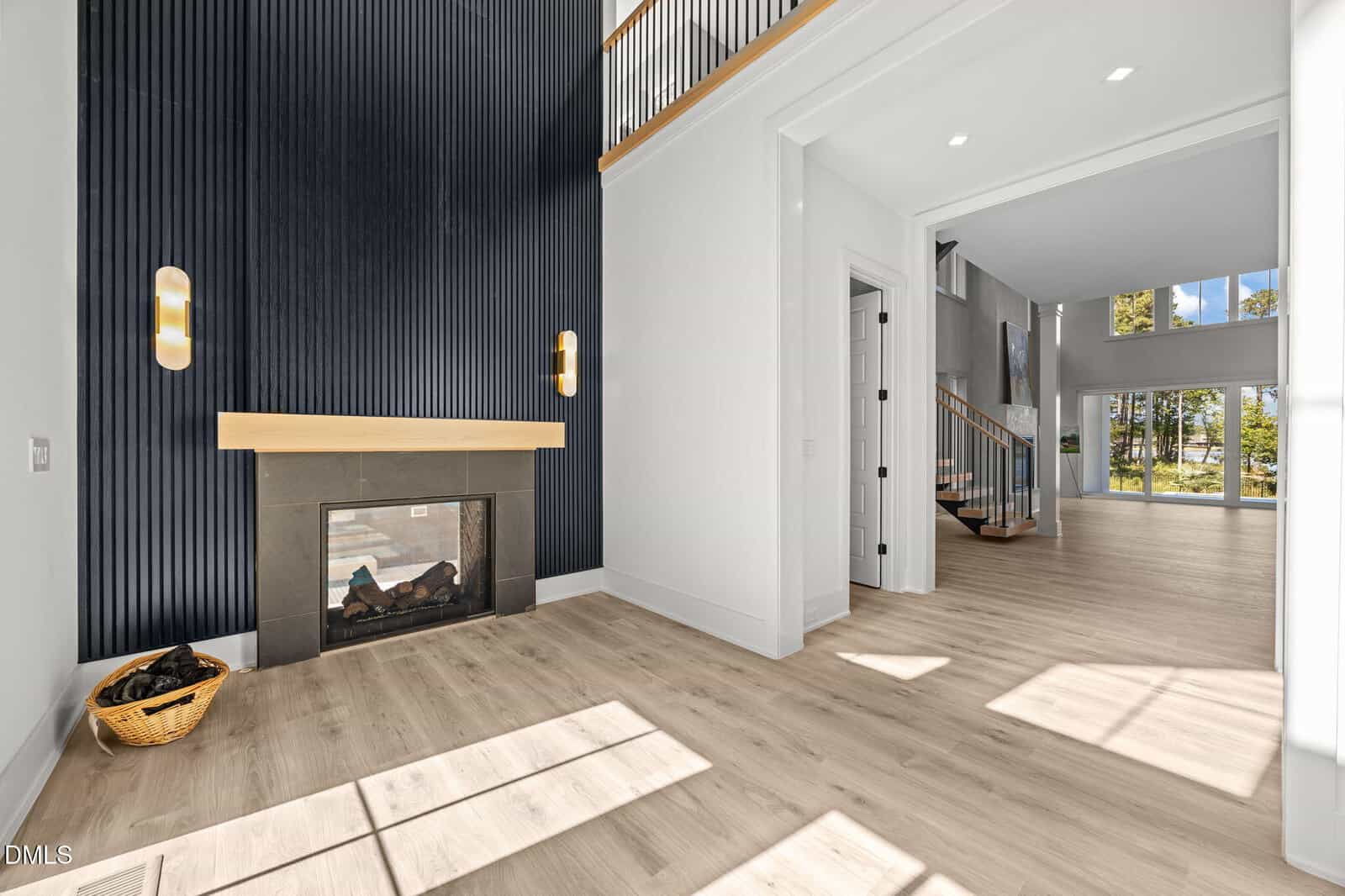 A modern, bright living space with light wood floors, a black fireplace with a wood mantel, black textured accent wall, two wall sconces, and large windows letting in natural light. A wicker basket of firewood sits by the fireplace.
