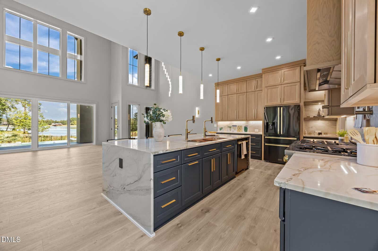 Modern kitchen with a large island, pendant lights, dark lower cabinets, light wood upper cabinets, marble countertops, and large windows overlooking a scenic lake view. Open floor plan with light wood flooring.