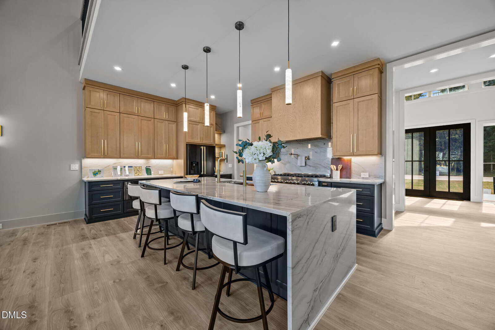 Modern kitchen with light wood cabinets, marble countertops, a large island with four upholstered barstools, pendant lights, and stainless steel appliances. Wide plank wood floors and double glass doors lead to the outside.