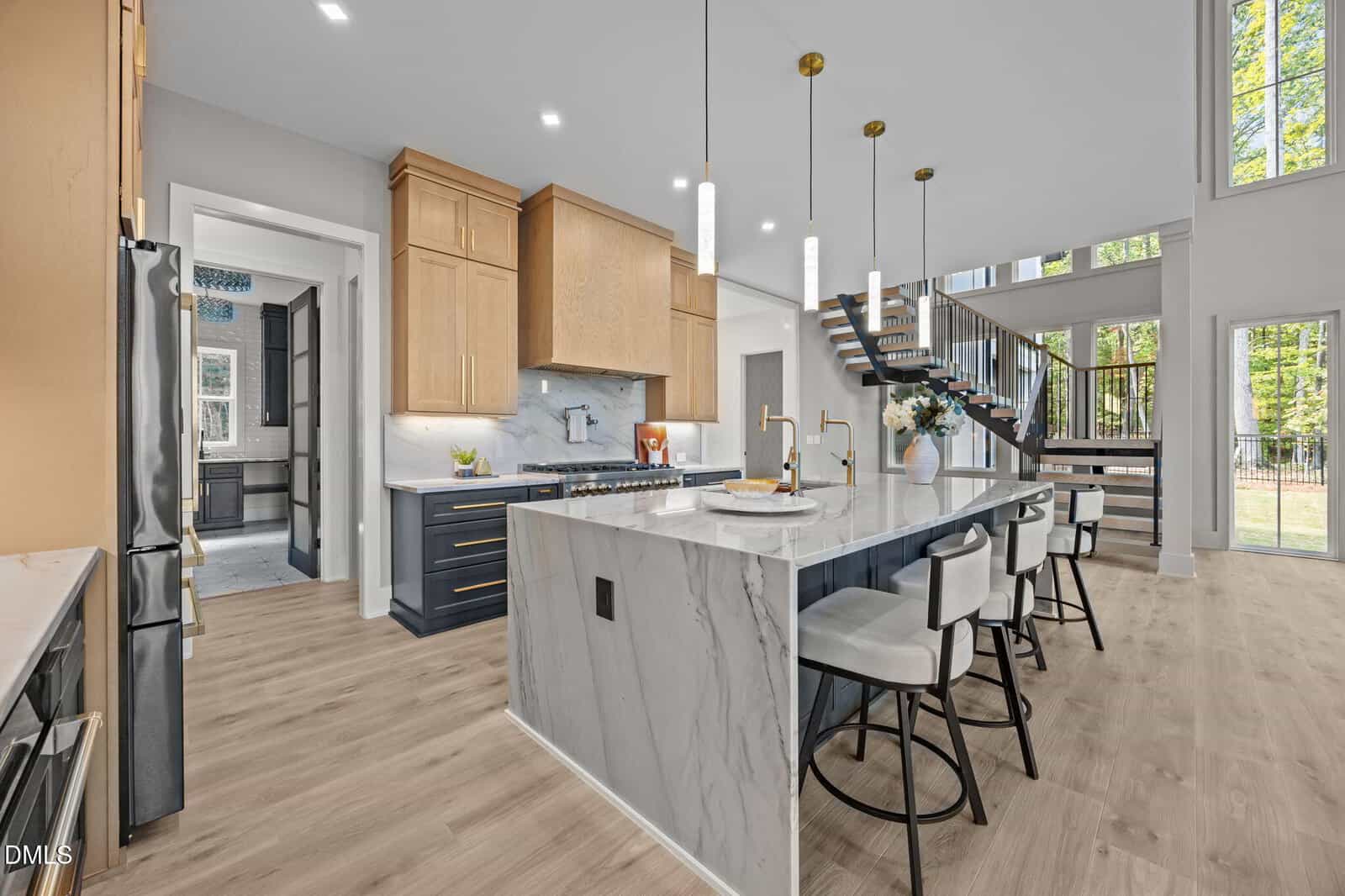 Modern kitchen with light wood floors, a large marble island with bar stools, gold pendant lights, wood and navy cabinets, and a floating staircase leading to a bright, open second floor with large windows.