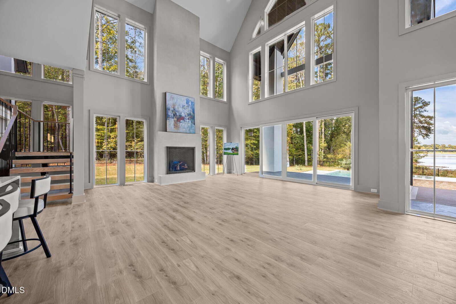 Spacious, modern living room with tall windows, light wood floors, a fireplace, and a staircase. Large windows reveal views of trees, a pool, and a nearby lake.