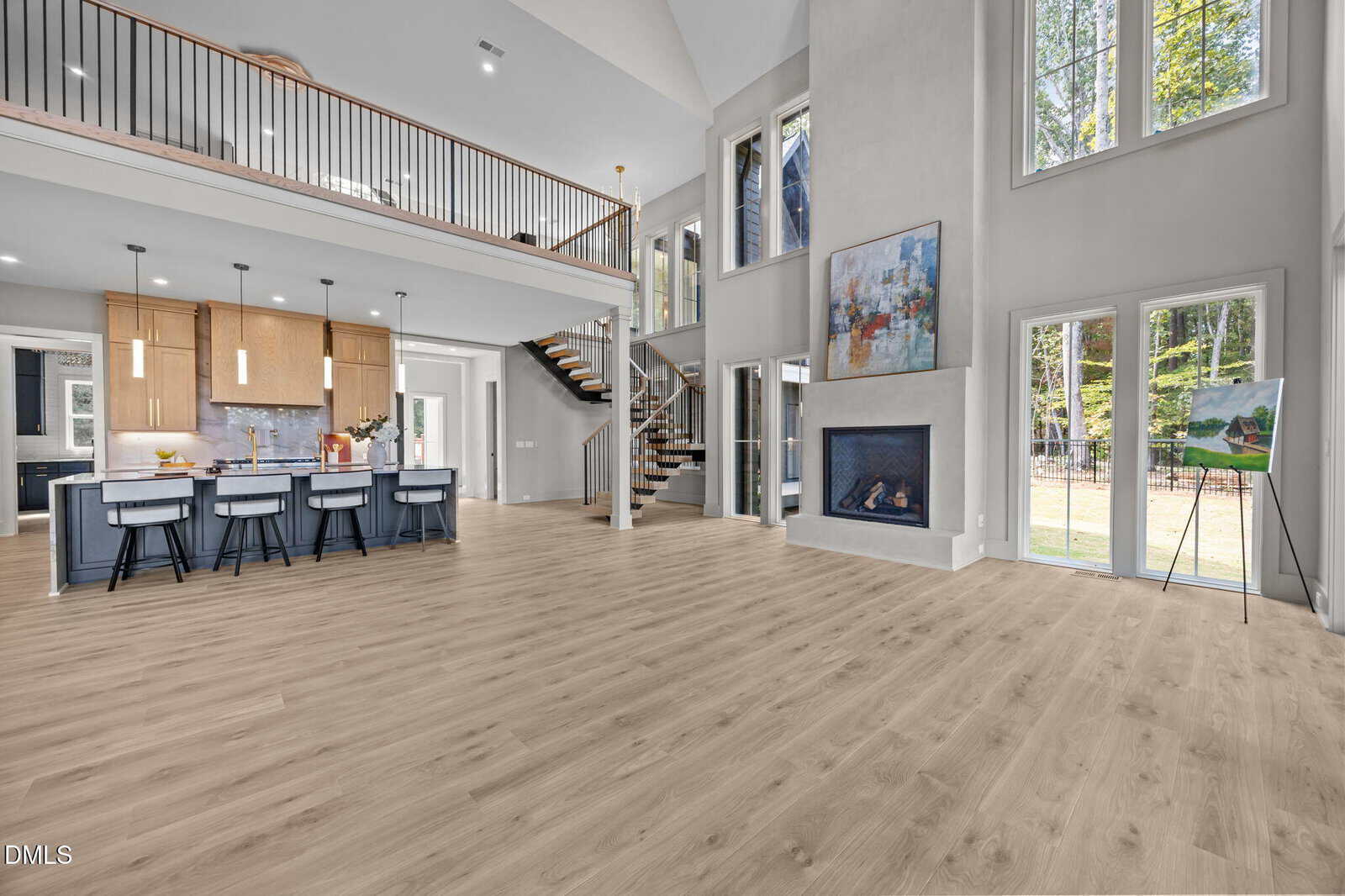 Spacious modern open-concept living room and kitchen with light wood floors, tall windows, a fireplace, wall art, and a staircase leading to a second-floor balcony railing.