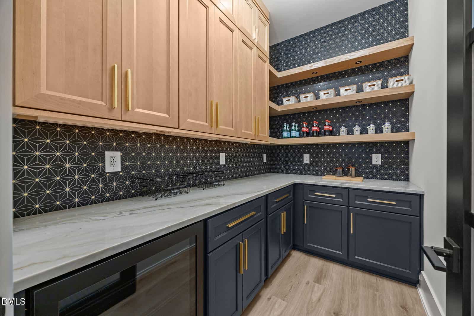 Modern pantry with light wood upper cabinets, dark blue lower cabinets, gold handles, and marble countertop. Open wooden shelves hold jars and spices against a geometric dark wallpaper. Light wood floor.