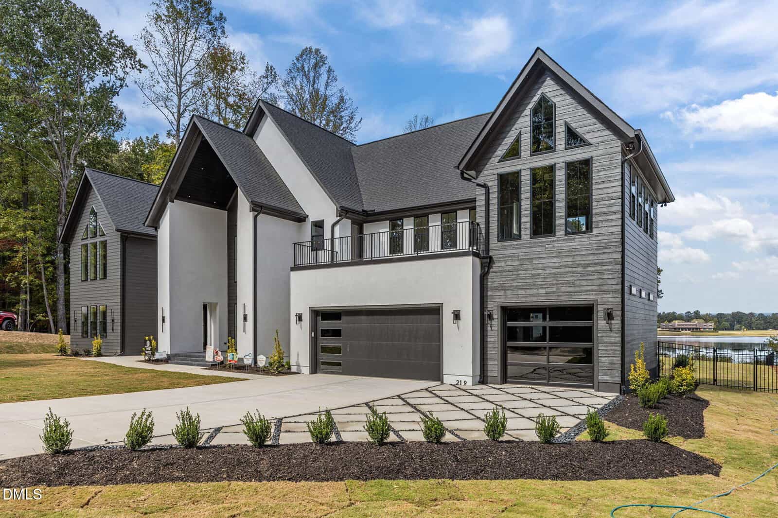 Modern two-story house with dark gray siding, large windows, and a three-car garage. The driveway features a geometric pattern, and the landscape is neatly manicured. Trees and a lake are visible in the background.