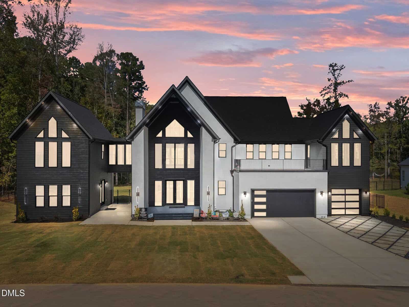 Modern two-story house at 219 Forest Bridge Road, Youngsville, featuring a black and white exterior, large windows, an attached three-car garage, and a spacious driveway, set against trees and a pink sunset sky.