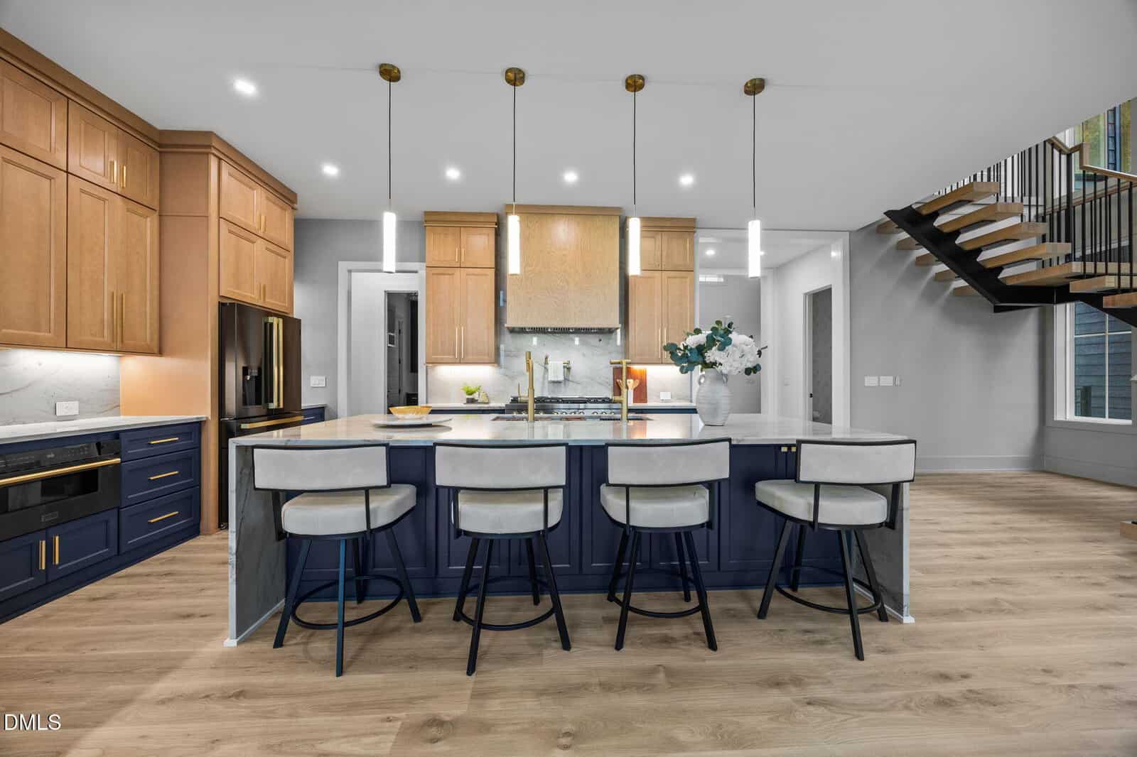 Modern kitchen with a blue island, four gray cushioned barstools, wood cabinets, pendant lights, built-in appliances, marble countertops, and a floating staircase. Light wood flooring and white walls create a bright, open space.