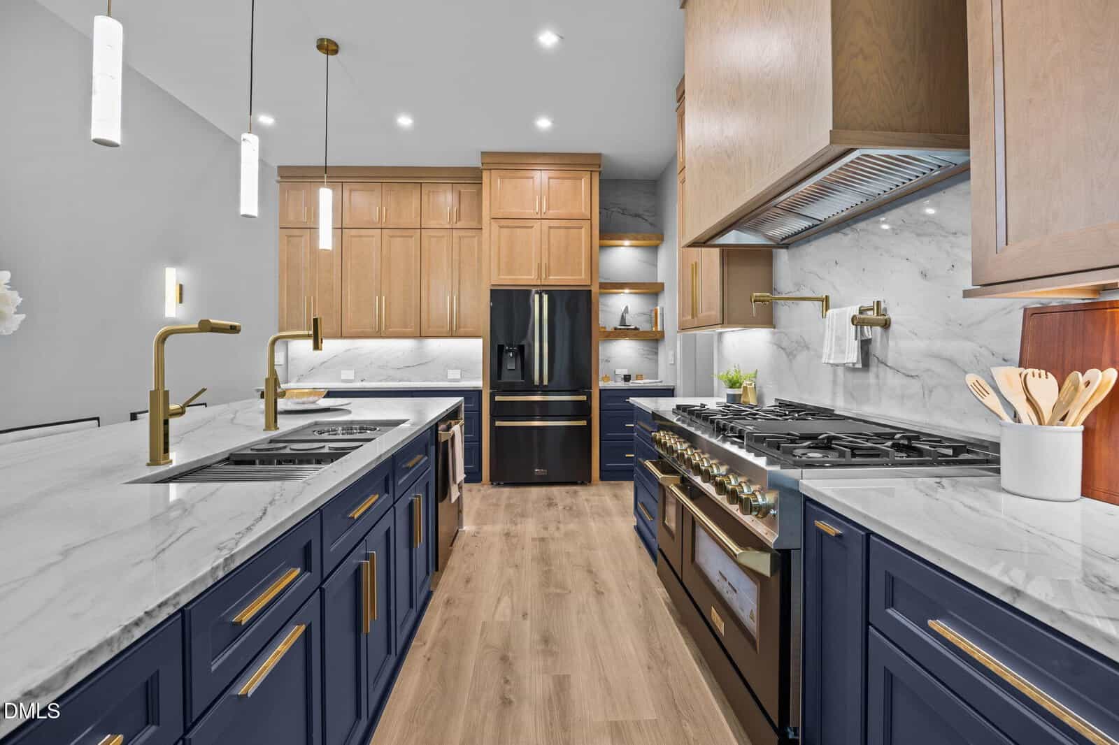 Modern kitchen with blue lower cabinets, gold hardware, marble countertops, wood upper cabinets, black refrigerator, double ovens, gas stove, and pendant lights, featuring a light wood floor and white marble backsplash.