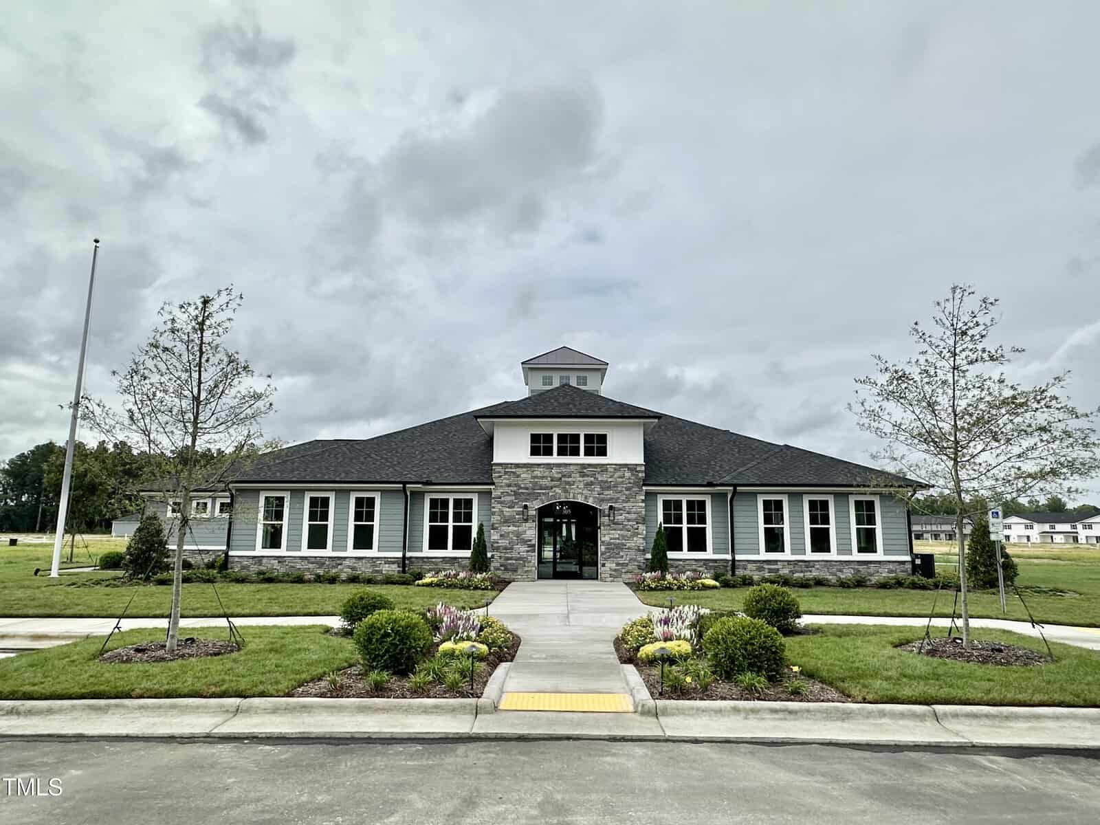 A one-story building with stone and gray siding, a central entrance, and a small cupola on the roof. The building is surrounded by neatly landscaped lawns, shrubs, and young trees under a cloudy sky.