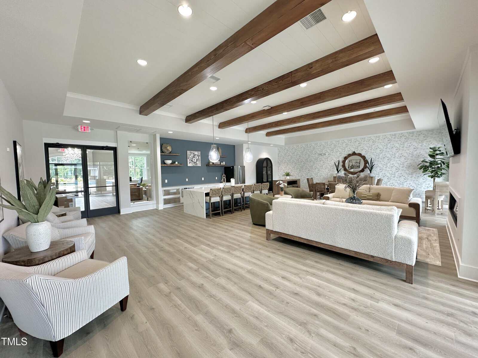 A spacious, modern lounge with wood beam ceiling, light wood flooring, neutral furniture, and a long communal table. Large windows and glass doors provide natural light. Decorative plants and wall art complete the decor.