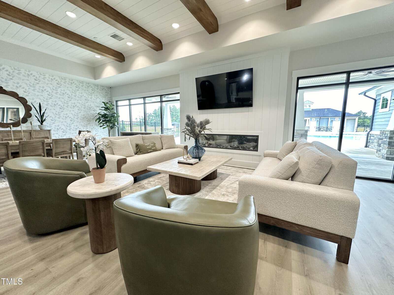 A modern living room with two light-colored sofas, two green armchairs, a coffee table, a wall-mounted TV above a linear fireplace, wood ceiling beams, large windows, and sliding glass doors opening to a patio.