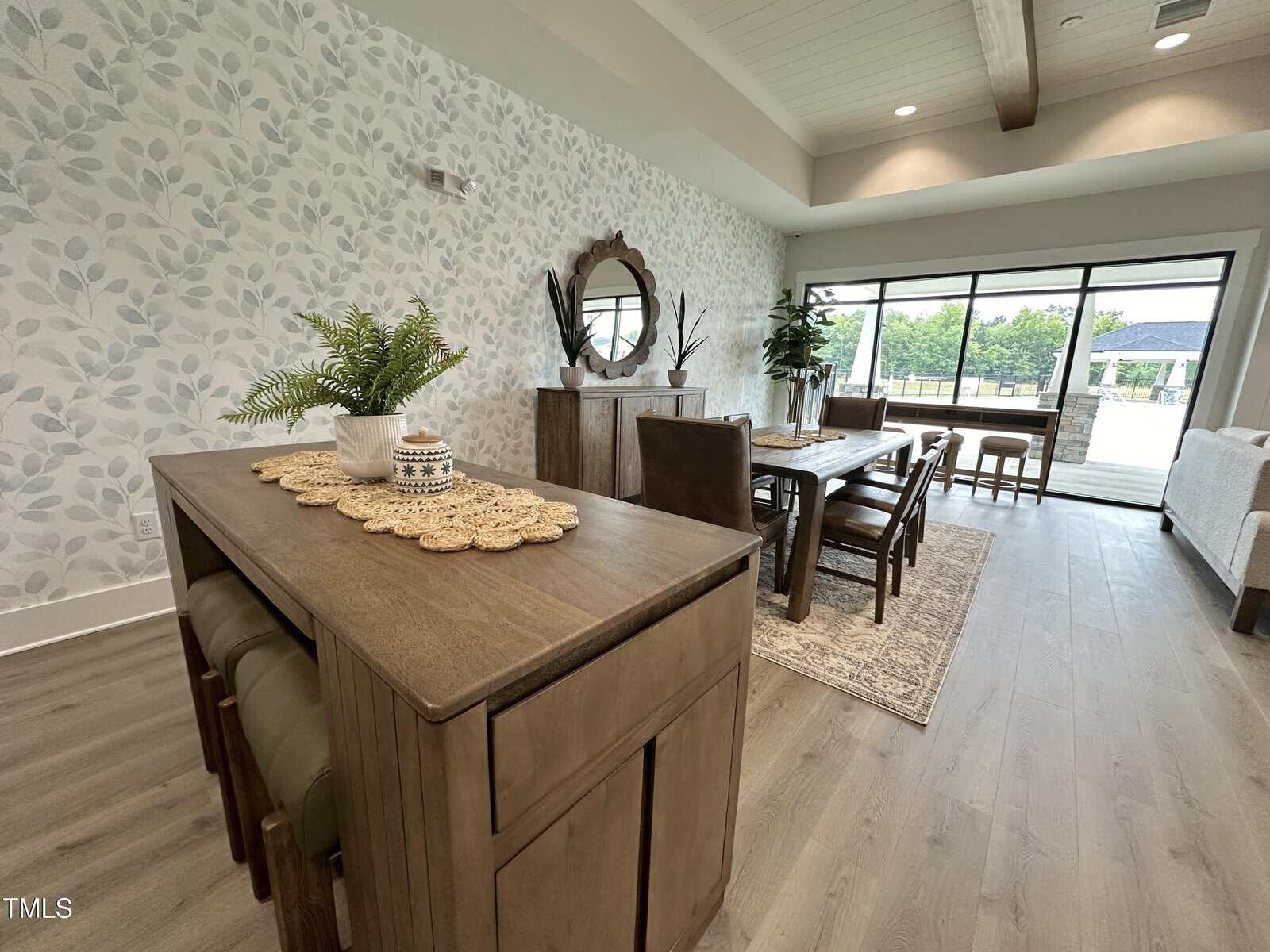 A dining area with a wooden table and six chairs on a patterned rug, a sideboard with a round mirror and decor, large windows with a patio view, and light wood flooring. A small plant sits on another wooden table in the foreground.
